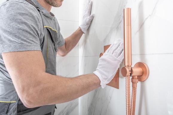 Plumber installing new matte rose gold shower fixtures on white marble tile wall.