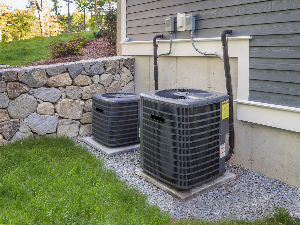 Two HVAC units installed outside a gray home, with green grass and stone wall behind them.