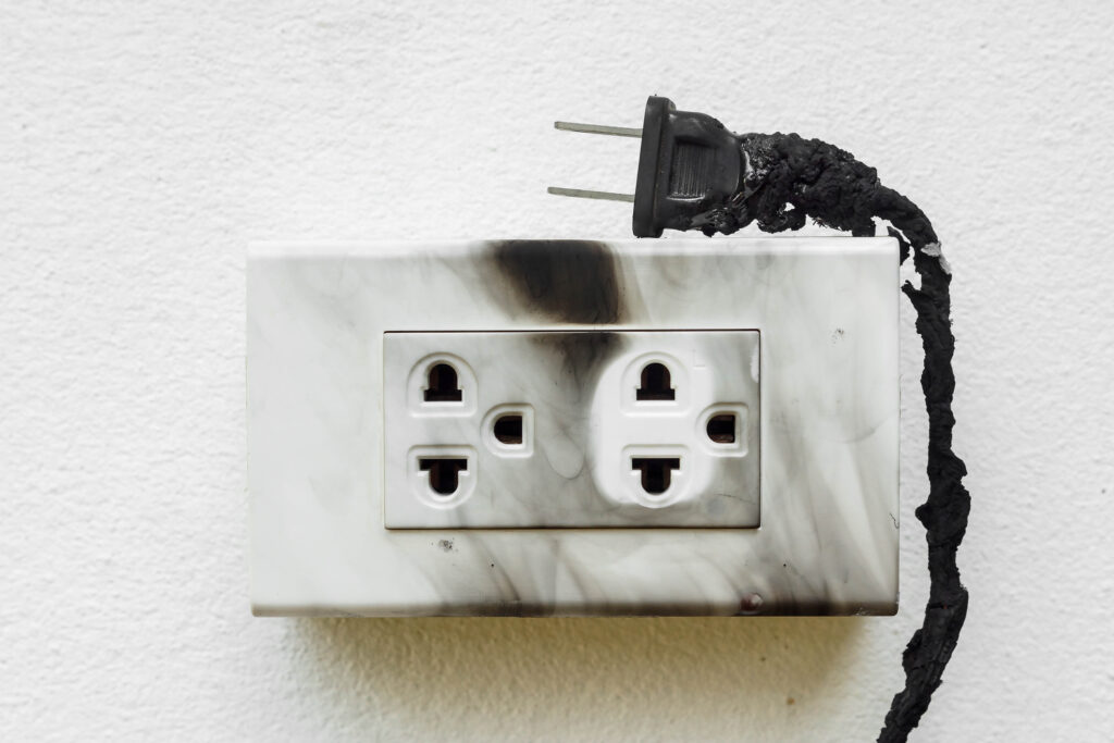 A wall outlet on a white wall, with burnt black electrical cord around it and scorch marks on the outlet cover.