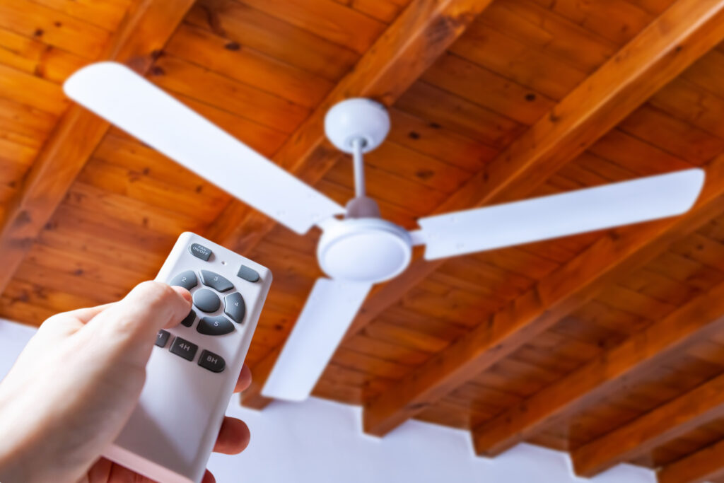 A hand using a remote control to operate a white three-blade ceiling fan mounted in a house on a wood beam ceiling.