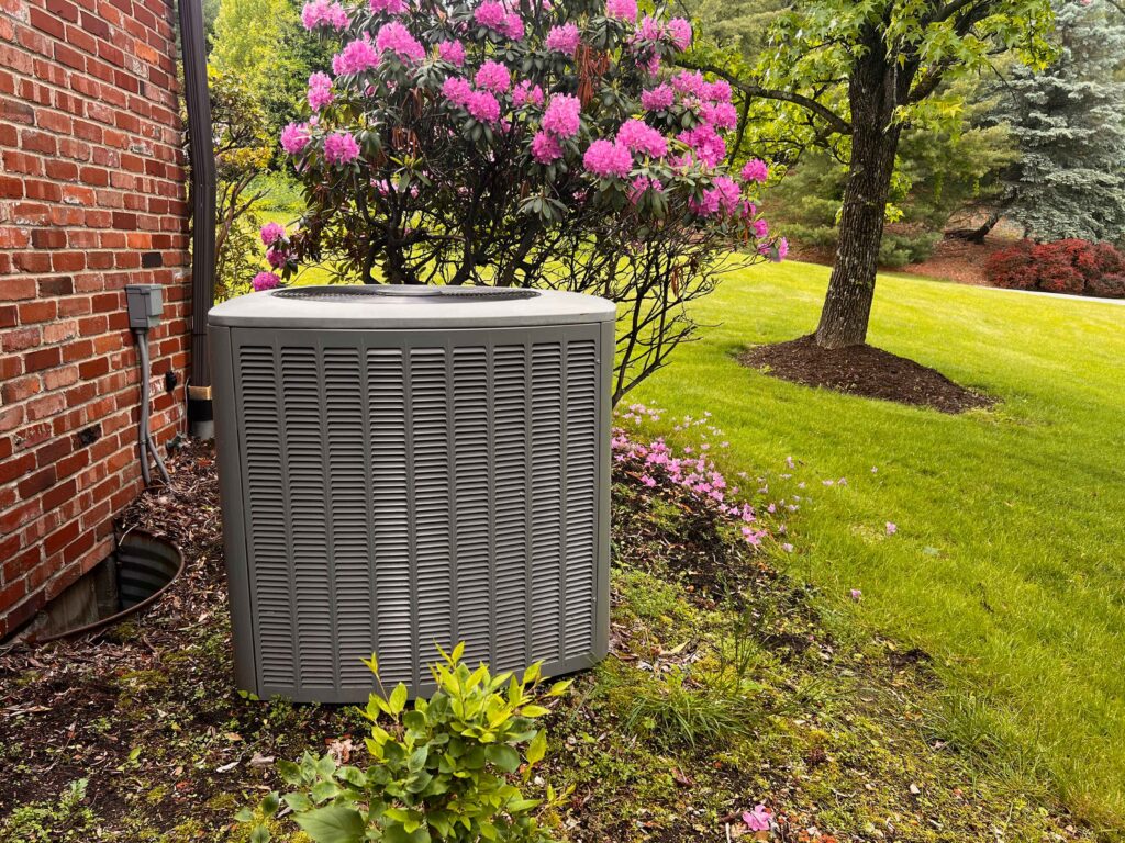 Air conditioner unit outside of a house with pink flowers surrounding it