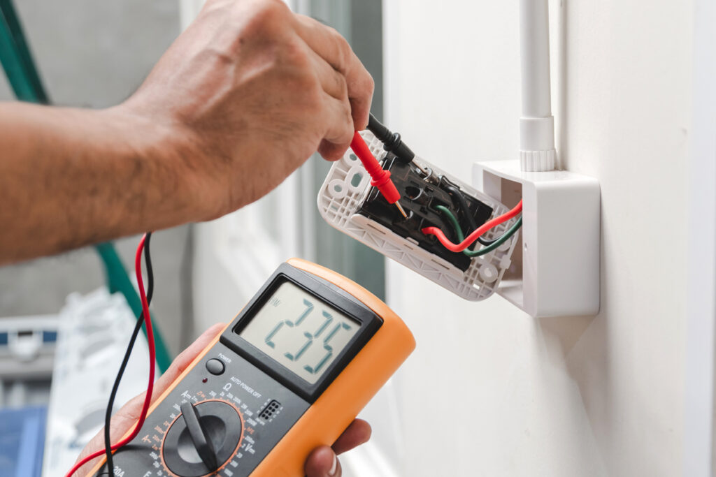 Electrician is using a digital meter to measure the voltage at the power outlet in on the wall.