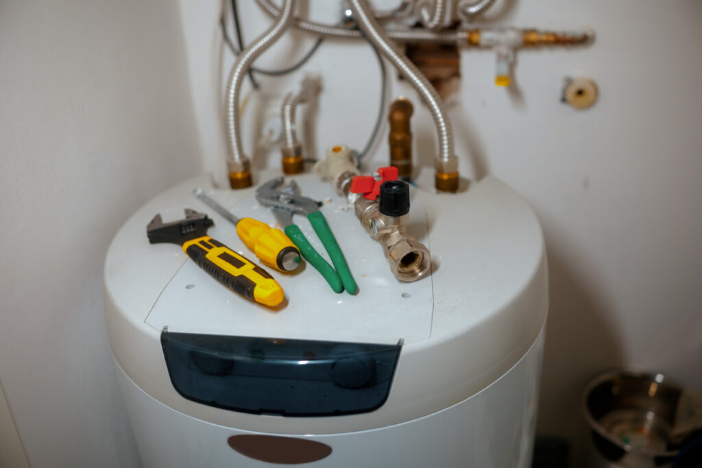 An adjustable wrench, screwdriver, and pliers rest on a white water heater, surrounded by pipes and valves, signaling active plumbing repair or maintenance work on a water heater